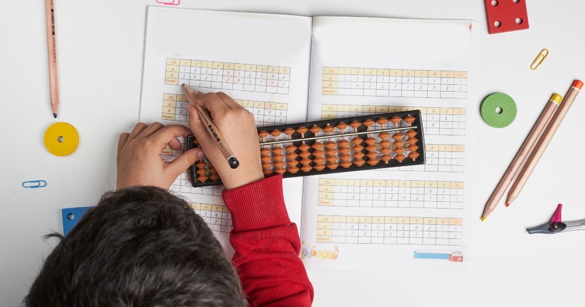 Child using an abacus to solve math problems in a workbook, surrounded by colorful pencils and learning tools. The image represents hands-on and visual learning techniques in Waldorf math education.